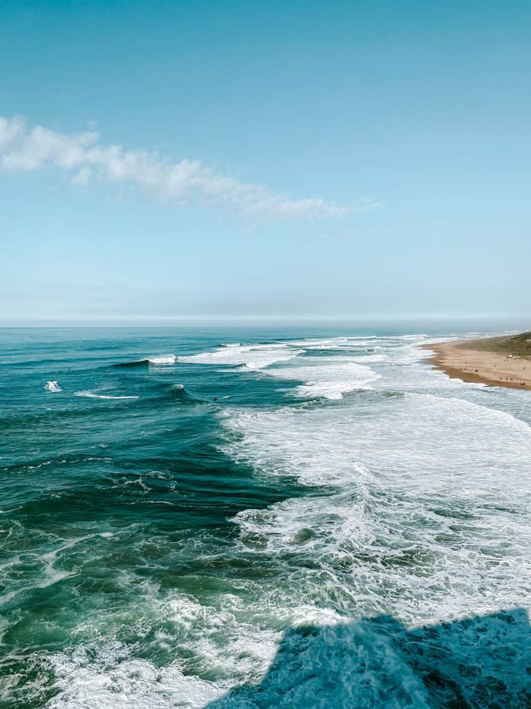 Uitzicht over de turquoise Atlantische Oceaan bij Nazaré waar lange rijen golven met wit schuim richting het uitgestrekte zandstrand rollen. Onder de helderblauwe lucht zijn enkele surfers als kleine stipjes tussen de golven te zien.