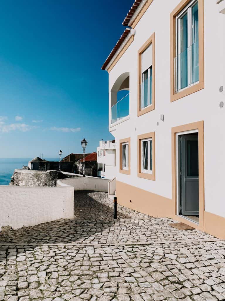 Smal straatje met kinderkopjes en witte huizen met zandkleurige details in het historische centrum van Nazaré. Het straatje loopt richting zee en laat de charmante Portugese architectuur zien die je tegenkomt tijdens het ontdekken van de bezienswaardigheden in Nazaré.