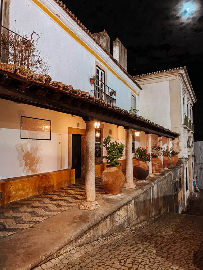 Rua de calçada portuguesa em Óbidos à noite, com uma varanda tradicional de colunas de pedra, vasos de barro com plantas e casas brancas com detalhes amarelos. Os candeeiros iluminam a arquitetura histórica da vila, sob um céu escuro com a lua parcialmente escondida pelas nuvens.